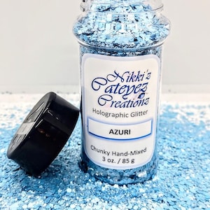 Puede incluir: Un frasco transparente lleno de purpurina holográfica azul y blanca gruesa. La etiqueta dice "Nikki's Crafty Creations", "Purpurina holográfica", "AZURI", "Grueso hecho a mano", y "3 oz. / 85 g".