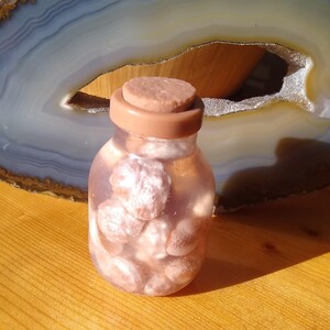 May include: A small glass jar with a cork lid filled with pink and white seashells. The jar is sitting on a wooden surface.