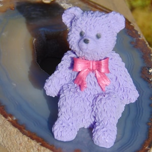 May include: A purple teddy bear with a pink bow sits on a slice of agate. The bear has a fuzzy texture and is sitting upright.