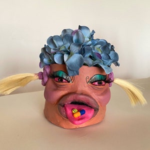 May include: A ceramic planter shaped like a face with blue flowers on top. The face has pink lips, green eyeshadow, and a tongue sticking out with two pills on it.