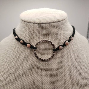 以下が含まれることがあります： ピンクと黒の斑点模様のビーズとシルバーの円形ペンダントが付いた、黒いチョーカーネックレスです。