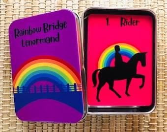 May include: A purple tin with the text "Rainbow Bridge Lenormand" and a rainbow design. Inside, a card with a black silhouette of a rider on a horse against a pink background, with a rainbow arching above the horse.