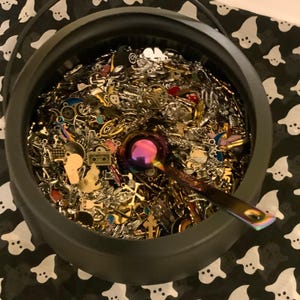 May include: A black cauldron filled with a variety of silver and gold charms, beads, and other jewelry findings. A purple metal spoon is resting on top of the charms.