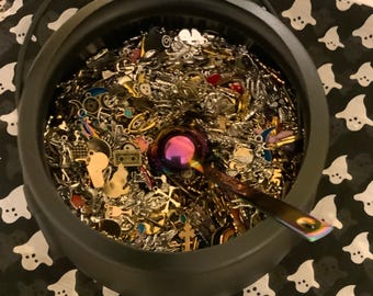 May include: A black cauldron filled with a variety of silver and gold charms, beads, and other jewelry findings. A purple metal spoon is resting on top of the charms.