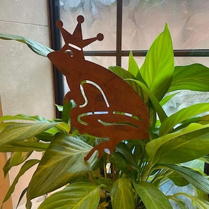 May include: A rusty metal frog with a crown on its head is sitting on a green plant in a pot. The frog is a garden decoration.