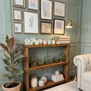 May include: A wooden shelf unit displays framed pictures, terrariums, clocks, books, and decorative jars. A potted plant and a brass floor lamp add to the room's decor. The wall is painted a muted green, creating a cosy and inviting atmosphere.