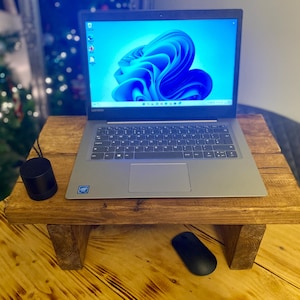 May include: A laptop computer rests on a rustic wooden laptop stand. The stand is stained a warm brown and has a simple, rectangular design with short legs. A black mouse and speaker are on the table. The laptop screen displays a blue and white abstract design.