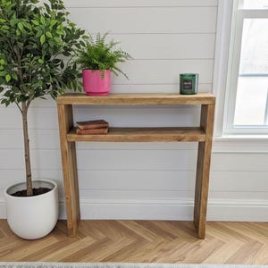May include: A wooden console table with a lower shelf, holding two small books. A pink potted plant and a green candle sit on top. A tall potted tree is to the left. The room has a white wall and a wooden floor.