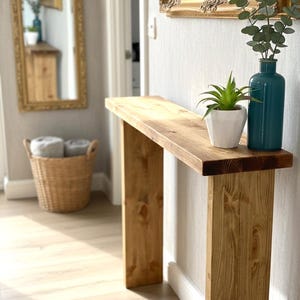 May include: A wooden console table with a natural finish. The table has a rectangular top and two square legs. A teal vase with greenery and a small potted plant sit on the table. A gold-framed mirror hangs on the wall. The table is approximately 120 cm long.