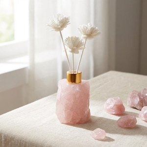 May include: A pink rose quartz diffuser with three white flower reeds. The diffuser has a gold-coloured collar. Several rose quartz stones are scattered on the table. The table is covered with a light-coloured fabric.