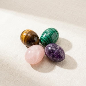 May include: Four polished gemstone eggs in a circular arrangement. The stones include tiger's eye, malachite, rose quartz, and amethyst. The eggs are smooth and glossy, with natural color variations.