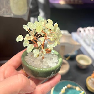 Könnte beinhalten: Eine kleine dekorative Baumskulptur mit einem Kupferdrahtstamm und hellgrünen Edelsteinblättern. Der Baum ist in einem kleinen, runden, grünen Keramiktopf mit weißen Steinen eingetopft. Die Skulptur wird in einer Hand gehalten.