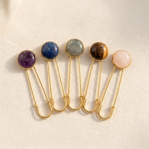 May include: Five gold-toned safety pins, each featuring a different circular gemstone topper. The stones include a deep purple amethyst, a dark blue lapis lazuli, a grey labradorite, a brown tiger's eye, and a pale pink rose quartz cabochon.