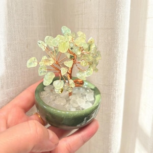 Op de afbeelding: Een kleine decoratieve boomsculptuur met een koperdraadstam en lichtgroene edelsteenbladeren. De boom staat in een kleine, ronde, groene keramische pot gevuld met witte stenen. De sculptuur is een miniatuur bonsai boom.
