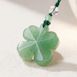 May include: A green aventurine clover pendant with a four-leaf design. The pendant is suspended from a dark green cord, accented with clear and green beads. The clover has a polished, translucent appearance.