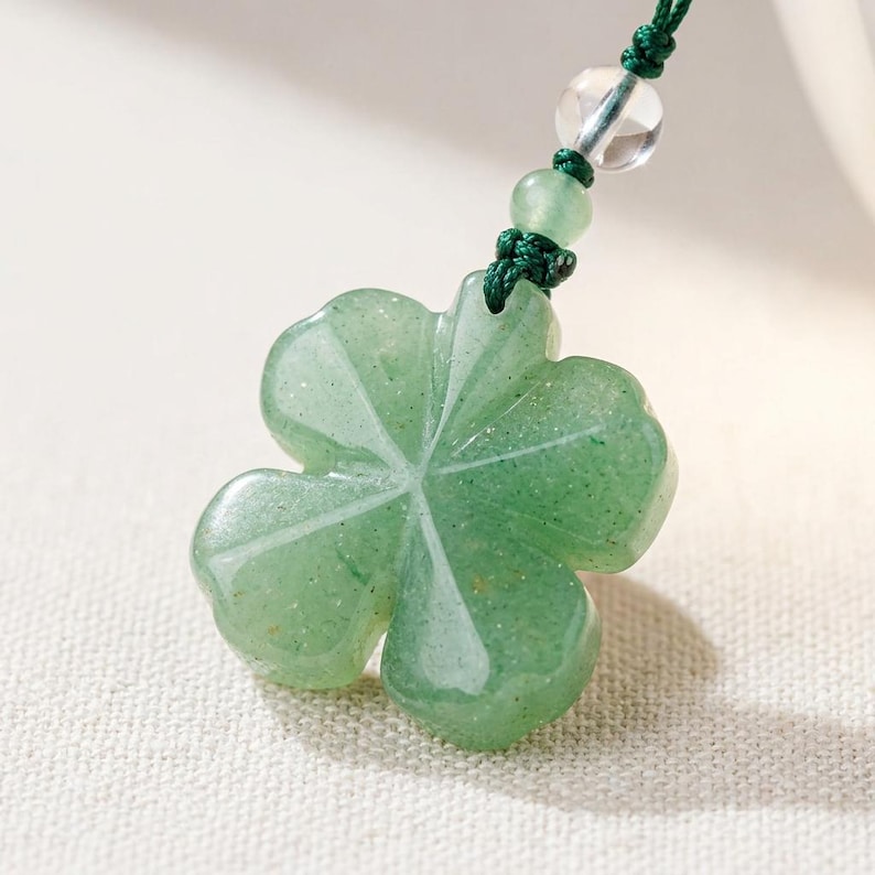 May include: A green aventurine clover pendant with a four-leaf design. The pendant is suspended from a dark green cord, accented with clear and green beads. The clover has a polished, translucent appearance.