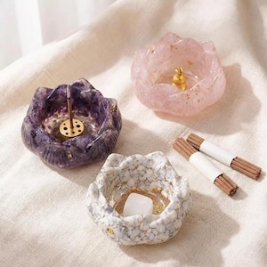 May include: Three lotus-shaped incense holders in purple, pink, and white with gold detailing. Incense sticks are bundled with white paper. The holders are made of a translucent material, ideal for home decor.