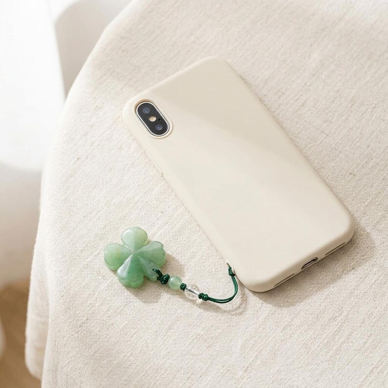 May include: A cream-colored phone case with a green four-leaf clover charm. The clover is attached to the case with a green cord and clear beads. The phone case is lying on a cream-colored fabric.