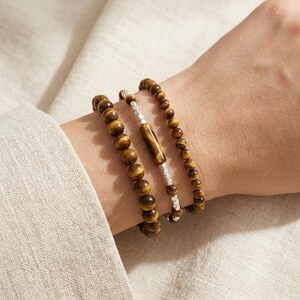 Puede incluir: Tres pulseras en una muñeca. Dos pulseras están hechas de cuentas redondas de ojo de tigre marrón. La tercera pulsera tiene cuentas de ojo de tigre, un cilindro de ojo de tigre y cuentas de color plateado. Las pulseras están apiladas juntas.