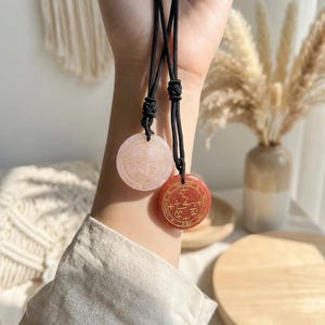 Op de afbeelding: Twee ronde hangerkettingen. Eén is lichtroze, de andere is rood-oranje. Beide hebben goudkleurige symbolen en tekst. De hangers hangen aan zwarte koorden. De achtergrond bevat gedroogde planten in een vaas en een houten oppervlak.