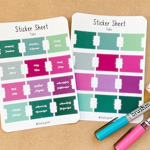 Könnte beinhalten: Zwei Stickerbögen mit monatlichen Registerkarten in verschiedenen Farben, darunter Grün, Rosa und Lila. Jede Registerkarte ist mit einem Monat in weißem Text beschriftet. Dazu zwei Brushmarker und eine Rolle Dekoband.