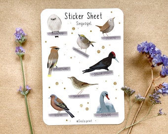 May include: A sticker sheet featuring illustrations of various birds, including a long-tailed tit, a wren, and a black woodpecker. The sheet has the text "Sticker Sheet Singvögel" and is decorated with gold dots. Lavender and blue flowers are in the background.