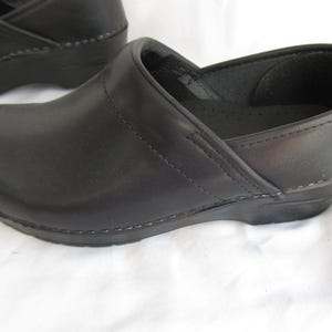 May include: A pair of black leather clogs featuring a rounded toe, a low heel, and visible tonal stitching along the side panels and sole. The interior of the shoe is lined with black material, and the outer surface has a smooth, matte finish.