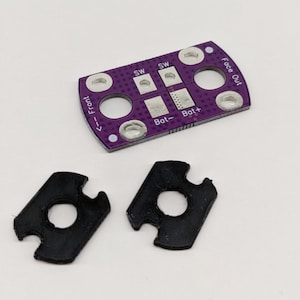 May include: A purple circuit board with white text that reads "Face Out", "Bot+", "Bot-", "SW", and "<-- Front". Two black rubber grommets are also shown.