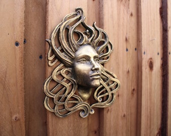 Könnte beinhalten: Eine goldfarbene Wandskulptur aus Metall, die das Gesicht einer Frau mit wallendem Haar darstellt. Die Skulptur ist an einer Holzwand befestigt.