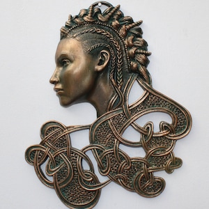 May include: A bronze wall sculpture of a woman's head with intricate Celtic knotwork. The woman has a braided hairstyle and a detailed facial expression. The sculpture is a rich brown color with green accents.
