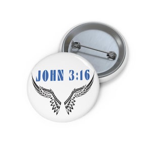 Puede incluir: Botón blanco con el texto "JOHN 3:16" en azul y un diseño de alas negras. Otro botón es visible en el fondo. Los botones son redondos y tienen un alfiler metálico en la parte posterior.