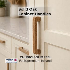 Può includere: Maniglie per mobili in legno di rovere massiccio con una sensazione robusta e solida. Le maniglie sono di colore marrone chiaro e sono attaccate a un'anta di un mobile color crema. Il testo "Solid Oak Cabinet Handles" è visualizzato nella parte superiore dell'immagine.