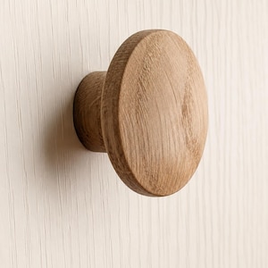 Puede incluir: Un pomo de mueble redondo de madera de color marrón claro con un patrón de veta natural visible. El pomo presenta una cara frontal circular plana y una base cilíndrica que se extiende hacia afuera, fijada a una superficie vertical de color blanco roto.