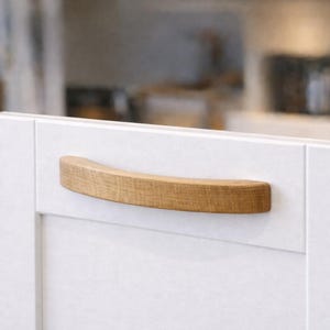 May include: A curved, light brown wooden cabinet handle with a visible natural grain pattern, mounted horizontally onto a flat, matte white cabinet door panel. The handle features a smooth, rounded profile and a solid, sturdy construction.