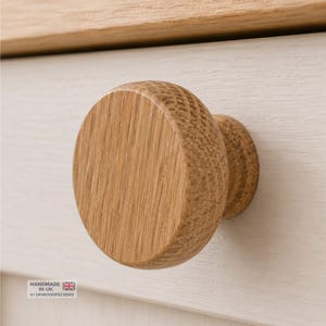 Op de afbeelding: Een ronde, lichtbruine houten meubelknop met een zichtbare houtnerf, bevestigd aan een wit geschilderd ladefront. De knop heeft een cilindrische basis en een plat, cirkelvormig oppervlak. Een label in de hoek vermeldt HANDMADE IN UK BY UKWOODPECKERS.