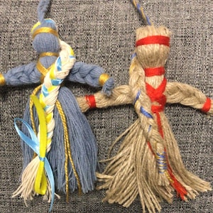 May include: Two handmade rag dolls with braided hair. One doll is blue with yellow and white braids and a blue ribbon. The other doll is brown with red and blue braids and a red ribbon.