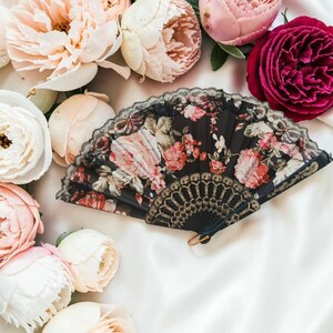 May include: A folding hand fan featuring a black fabric base with a vibrant floral print of pink and red roses. The fan is edged with black lace and includes a decorative gold-toned plastic handle with intricate cut-out patterns and a small ring.