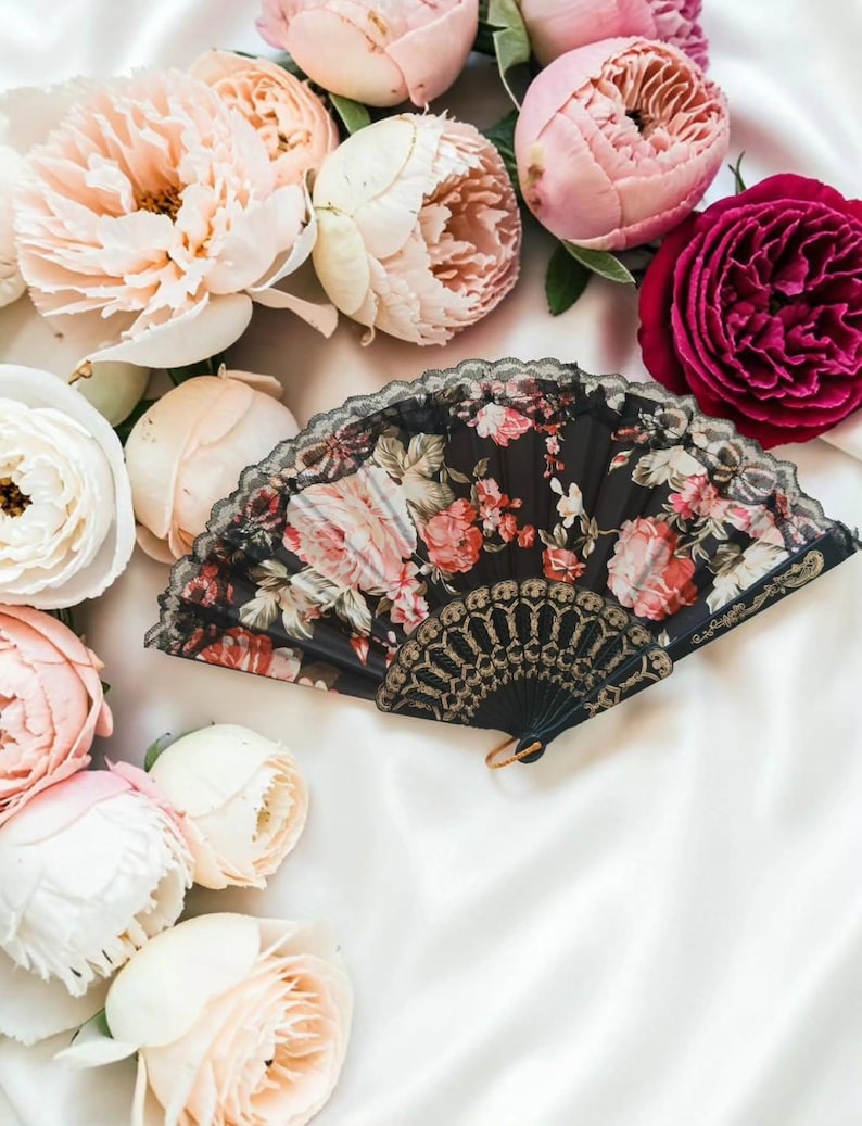 May include: A folding hand fan featuring a black fabric base with a vibrant floral print of pink and red roses. The fan is edged with black lace and includes a decorative gold-toned plastic handle with intricate cut-out patterns and a small ring.