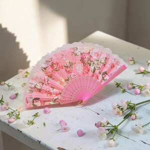 May include: A folding hand fan featuring a vibrant pink background with a detailed floral pattern of roses and green leaves. The fan has a delicate white lace trim along the top edge and a pink plastic frame with intricate cut-out detailing.