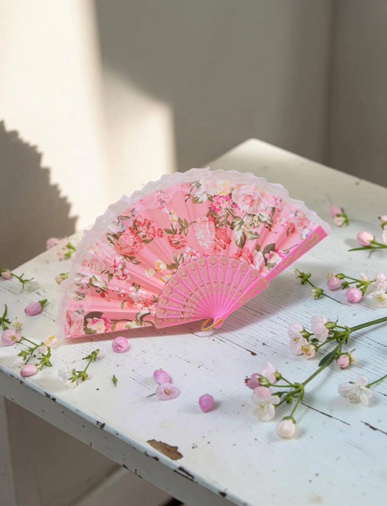 May include: A folding hand fan featuring a vibrant pink background with a detailed floral pattern of roses and green leaves. The fan has a delicate white lace trim along the top edge and a pink plastic frame with intricate cut-out detailing.