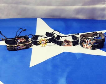 May include: Five leather bracelets with different charms, including a Puerto Rican flag, a sun, and a Puerto Rico logo. The bracelets are displayed on a blue and white fabric background.