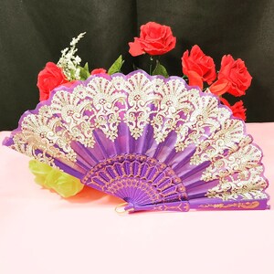May include: A purple folding hand fan featuring intricate gold-colored lace embroidery along the top edge. The fan has a solid purple handle and a series of decorative ribs that extend from the base to the fabric canopy, creating a pleated design.
