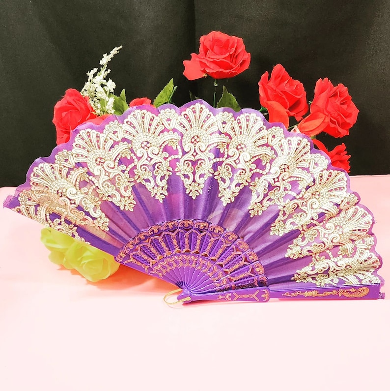 May include: A purple folding hand fan featuring intricate gold-colored lace embroidery along the top edge. The fan has a solid purple handle and a series of decorative ribs that extend from the base to the fabric canopy, creating a pleated design.