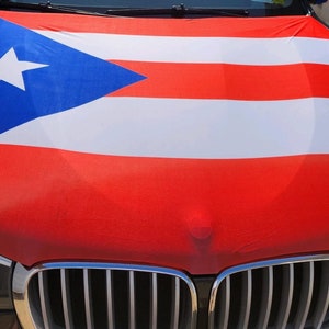 May include: A black car with a Puerto Rican flag covering the hood. The flag is red, white, and blue with a white star in the blue triangle.