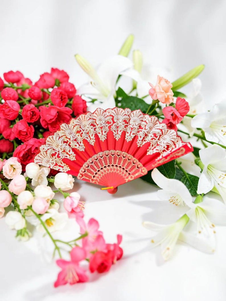May include: A vibrant red folding hand fan featuring intricate gold-colored lace detailing along the upper edge and a decorative pattern on the base, displayed amidst a cluster of white lilies and small pink and red rose-like flowers.