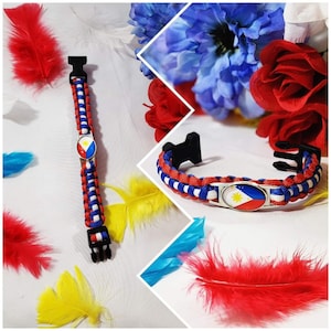 Könnte beinhalten: Ein rot-weiß-blaues Paracord-Armband mit einem Metallanhänger, der die Flagge der Philippinen zeigt. Das Armband ist vor einem weißen Hintergrund mit bunten Federn zu sehen.