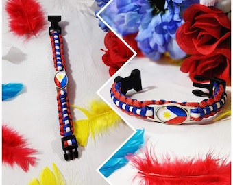 Puede incluir: Una pulsera de paracord roja, blanca y azul con un dije de metal que presenta la bandera de Filipinas. La pulsera se muestra sobre un fondo blanco con plumas de colores.