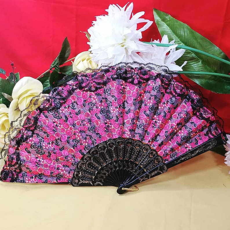 May include: A folding hand fan featuring a vibrant pink and purple floral pattern on a fabric surface. The top edge is trimmed with black lace, and the handle is constructed from black plastic with intricate cut-out detailing and gold accents.