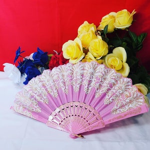 May include: A pink folding hand fan featuring intricate white lace overlays with gold accents along the top edge. The base of the fan is made of pink plastic with gold-colored filigree detailing. The fan is displayed in a semi-open position.