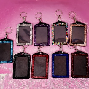 Puede incluir: Una colección de llaveros rectangulares con interior negro y borde decorativo de brillantes de colores. Los llaveros vienen en varios colores, incluyendo azul, plateado, morado y rojo, y están unidos a llaveros plateados.